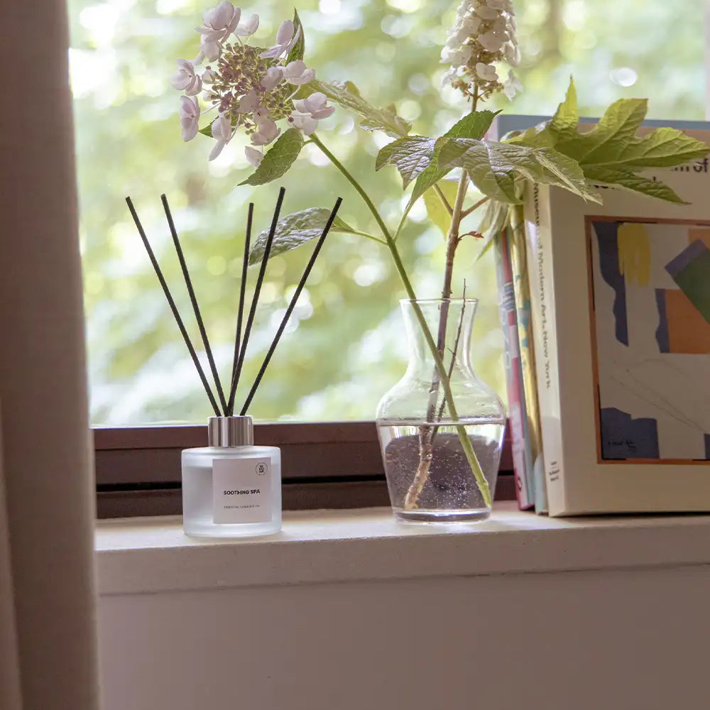 Doftpinnar med doften Soothing Spa ståendes i fönster tillsammans med blomma 