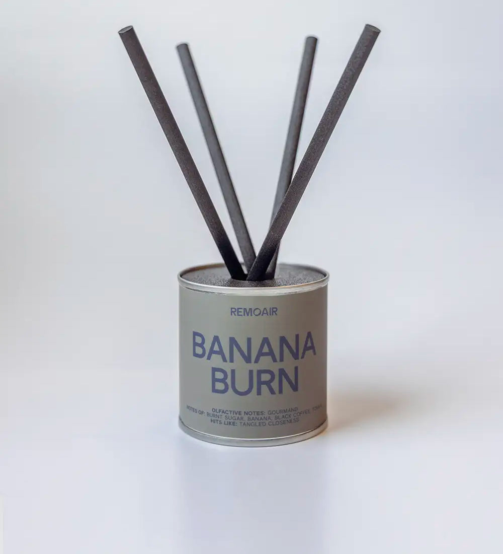 Banana Burn doftpinnar i metallburk med svarta stickor ⎯ ren neutral bakgrund i mjukt ljus.