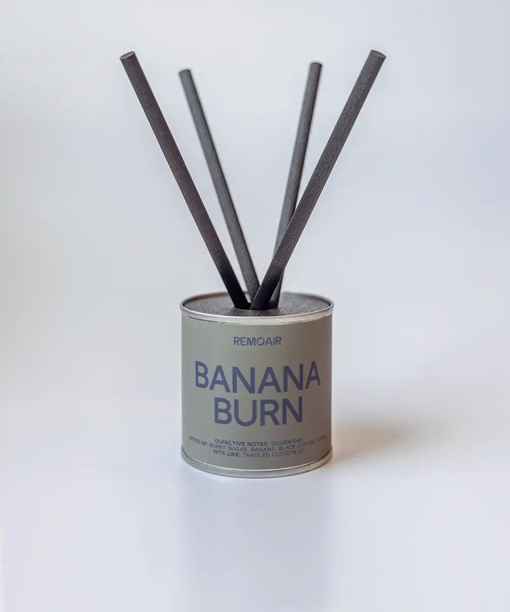 Banana Burn doftpinnar i metallburk med svarta stickor ⎯ ren neutral bakgrund i mjukt ljus.