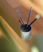 Astral Buds doftpinnar i silver plåtburk på ljust parkettgolv i varmt solljus.